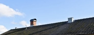 Svart tegeltak med skorsten och två ventilationstrummor mot en klarblå himmel.