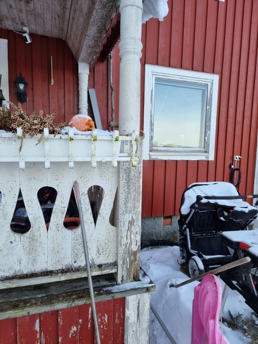 Vintertagen fasad på ett hus med skadad vit stupränna och fönster, sliten färg och snötyngd veranda.