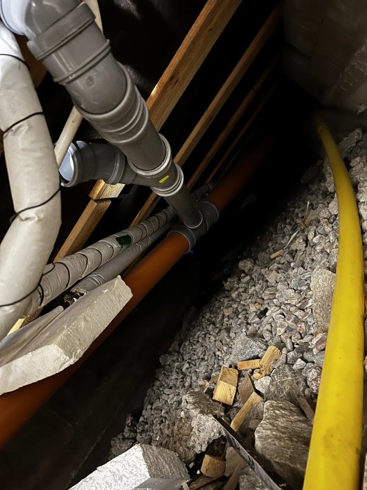 Plaströr och kablar i en torpargrund med synligt öppet avloppsrör och grusunderlag.