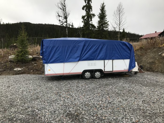 Husvagn täckt med blå presenning parkerad på grus på byggtomt omgiven av skog.