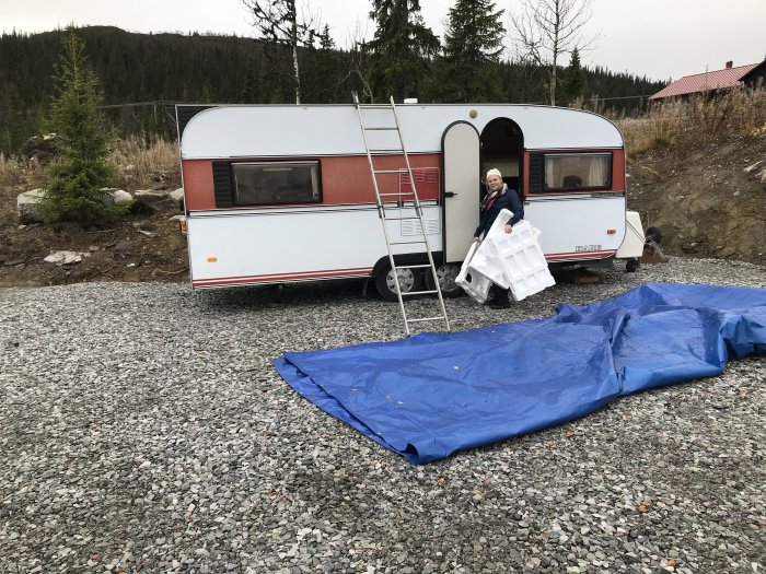 Husvagn parkerad på grusig tomt med person som bär stolar, skog i bakgrunden.