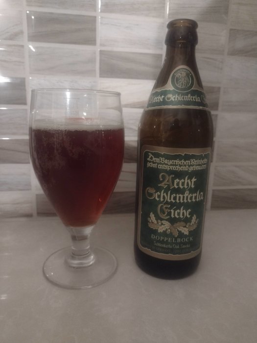 Ett glas fyllt med rödbrun öl bredvid en flaska märkt "Aecht Schlenkerla Eiche Doppelbock" på ett marmorbord.