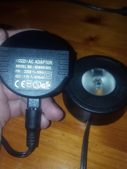 Hand som håller en AC-adapter bredvid en lampa med synlig glödlampa på ett träbord.