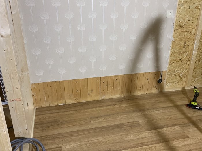 Inre hörn av ett rum med halvfärdig vägg, OSB-skiva nertill och tapetserad vägg ovanför, med synlig golvlist.