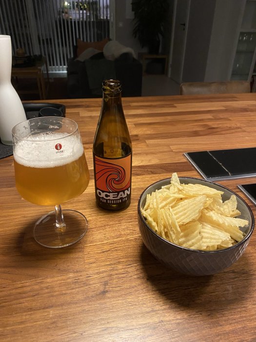 Ölglas med skummad öl bredvid öppen flaska märkt Ocean, skål med potatischips på träbord, inomhusmiljö i bakgrunden.