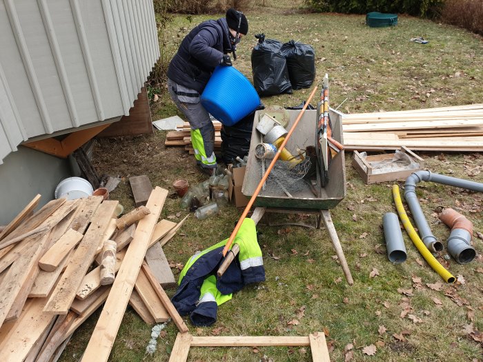 Två personer rensar röran av diverse byggmaterial och gamla saker utanför ett hus.