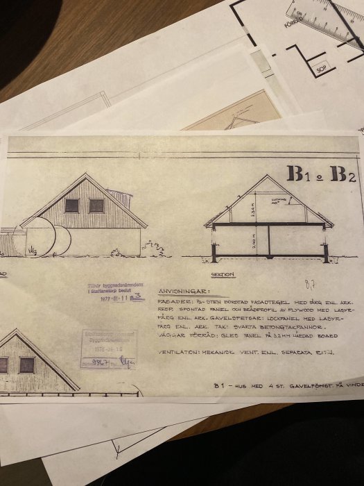 Gammal arkitektritning av ett 70-tals hus visar fasader och sektion, ligger på ett bord med andra ritningar.