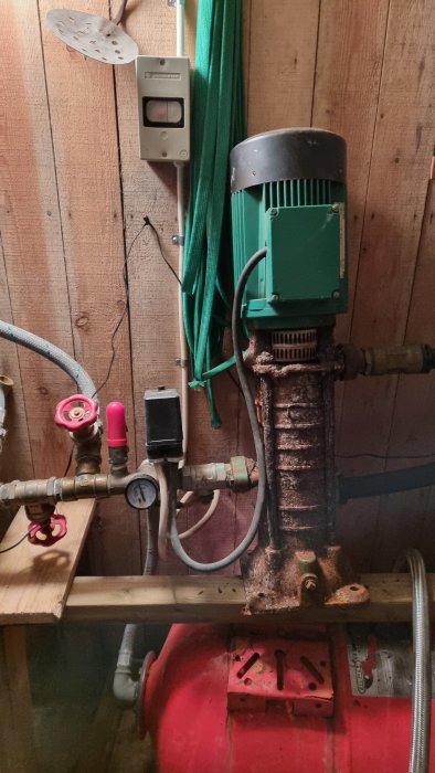 Rostig pump ovanpå röd hydrofor, med diverse rörkopplingar och grön slang bredvid en energimätare.