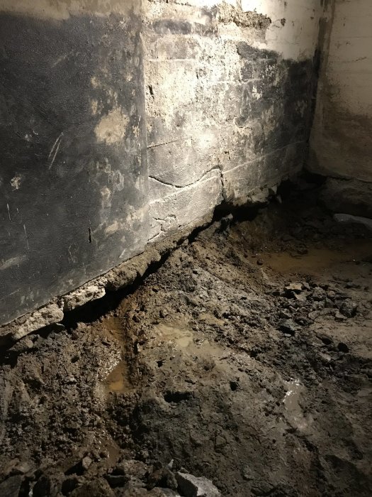 Uppgrävt och delvis spräckt betonggolv i källare med synlig berggrund och fukt vid väggens bas.