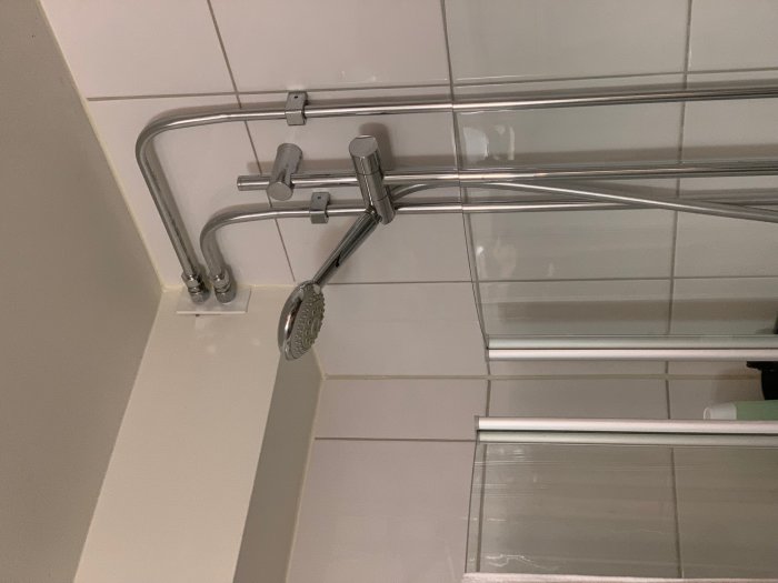 Kromade radiatorrör längs vägg och tak i ett badrum med duschhuvud och kakel.