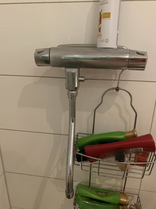 Äldre silverfärgad badrumsblandare på vägg ovanför en duschhylla med flaskor, söker reservdel.