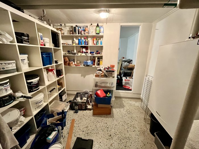 Två angränsande rum fulla av byggmaterial och verktyg, med hyllor och oorganiserade objekt på golvet.
