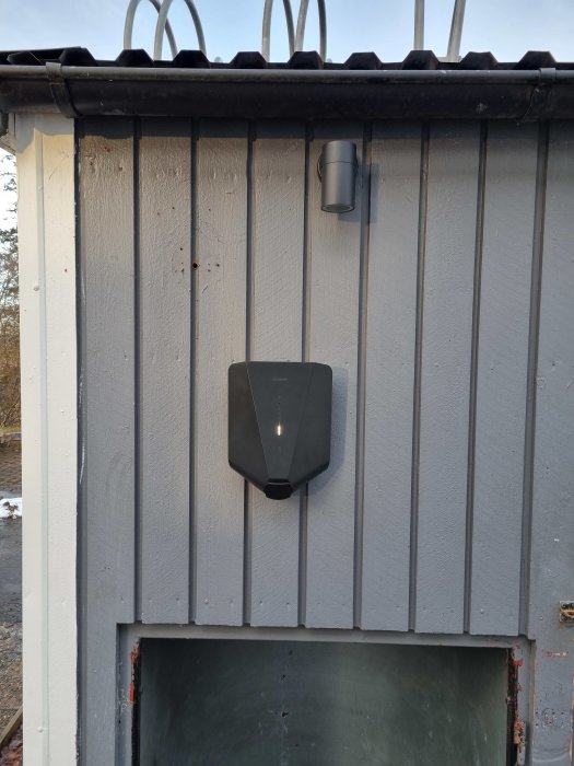 Laddbox installerad på gråmålad förrådsvägg ej centrerad under en svart utomhuslampa, med förberedelser för framtida ombyggnation.