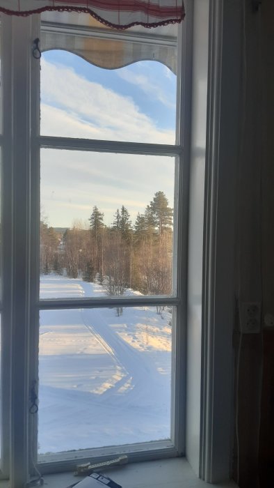 Högt treglasfönster inifrån ett hus med utsikt över ett snötäckt landskap och träd.