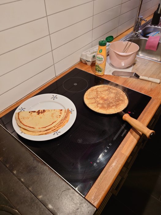 Pannkakor på spisen med stekspade, smörflaska, äggkartong och bunke på en köksbänk.