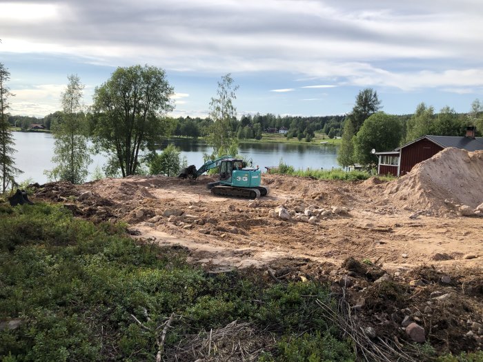 Tomt med pågående markarbete och grävmaskin, vid sjön, i förberedelse för husgrund.