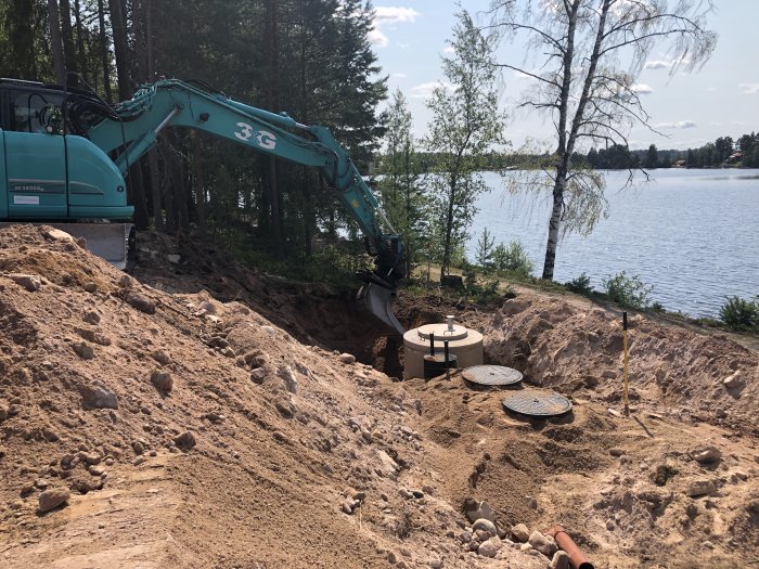 Grävmaskin vid sjö som förbereder grundarbeten för husbygge; grävningsarbete nära vatten med synlig minireningsverk.