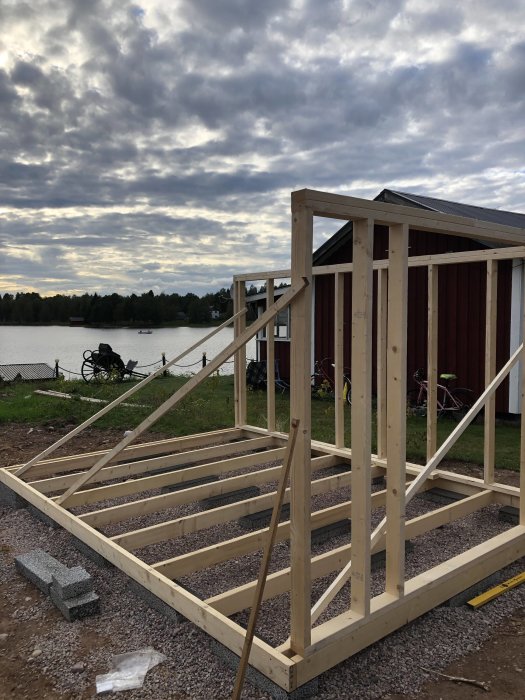 Grundläggning av en friggebod med trästomme under uppbyggnad framför sjö och molnig himmel.