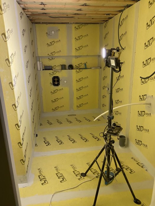 Badrum under renovering med gula LIP isoleringsskivor på väggar och golv samt ett stativ med belysning.