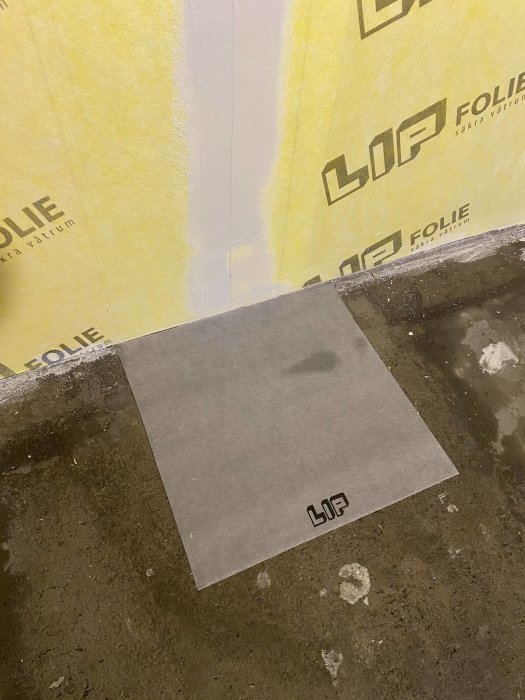 Byggisolering med LIP-logotyp på vägg och åtskild brunsmarchett på betonggolv vid väggkant.