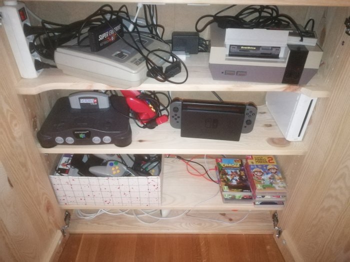 Hylla med olika spelkonsoler och spel, inklusive Nintendo 64, Switch och två staplade spel i förgrunden.