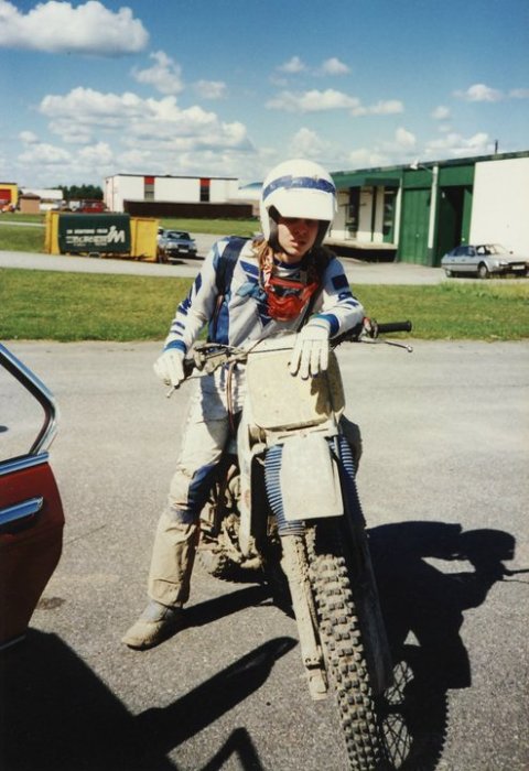 Ung person i crossutrustning står bredvid en lerig motocrosscykel, fotograferad utomhus för cirka 35 år sedan.