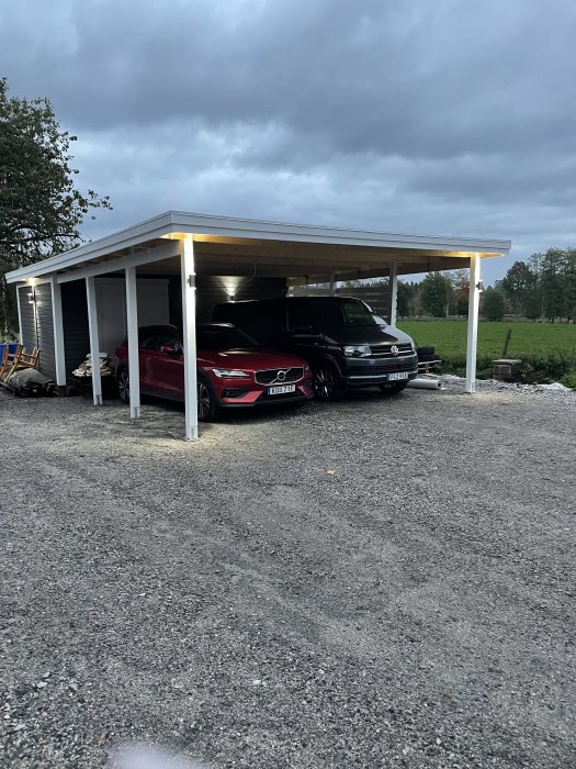 Nybyggd vit carport med plats för två bilar, grusgolv och belysning, omgiven av grönska.