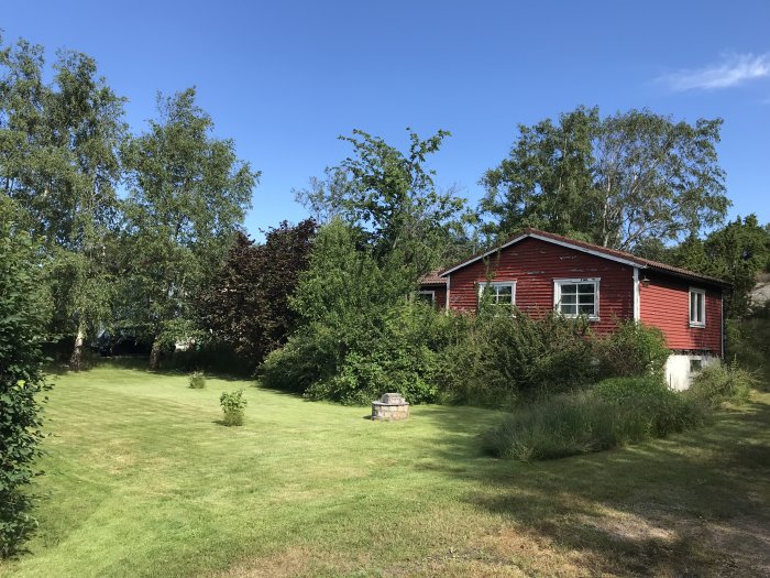 Röd trästuga med vitmålade fönster omgiven av en frodig trädgård och tät vegetation under en klarblå himmel.