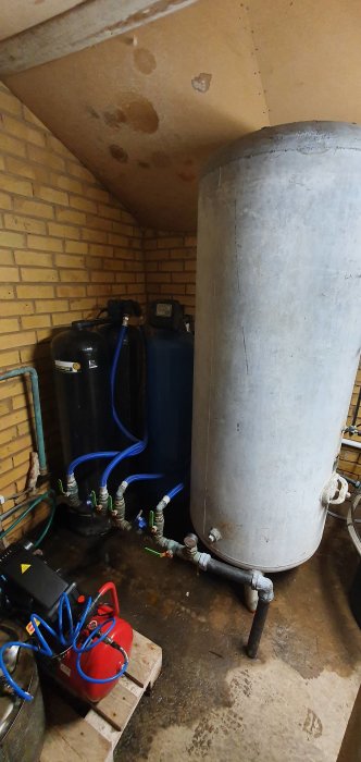 Gammal hydrofor och tryckmätare i ett pannrum med flera filter och en rörmotorpump.