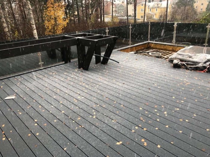 Terrass med glasräcke och förberedd plats för jacuzzi under snöfall, omgiven av höstlöv.