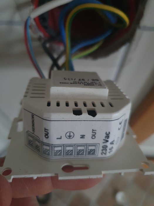 Hand håller en digital termostat med oanslutna färgglada ledningar i bakgrunden.
