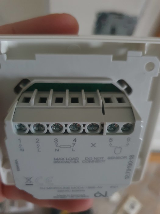 Närbild av en hand som håller en digital termostat med okopplade terminaler och ledningsdiagram.