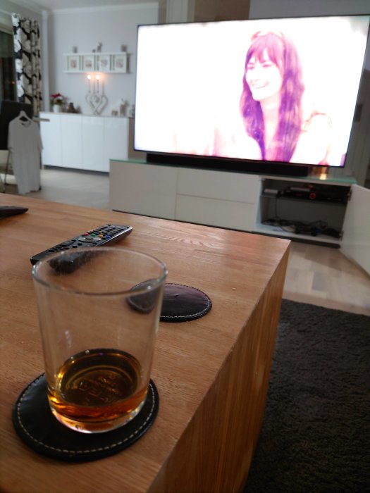 Glas med dryck på bord framför tv-bänk med påslagen TV och suddig bakgrund av ett vardagsrum.