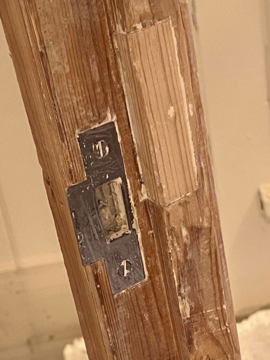 Delvis skrapad träyta på en dörrkarm med synliga spår av ett gammalt kammarlås, under lager av gammal färg.
