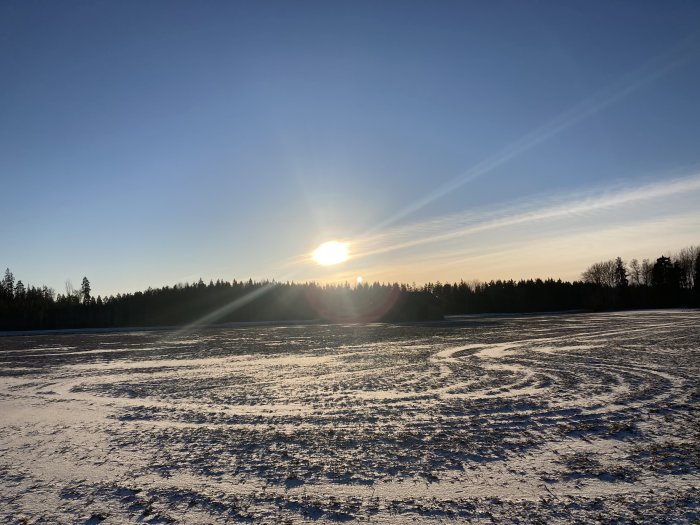 Solnedgång över ett snötäckt fält med hjulspår och skog i bakgrunden.