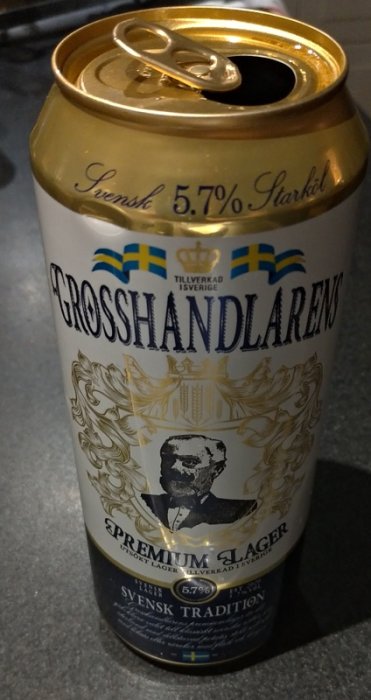 Öppnad ölburk av "GROSSHANDLARENS Premium Lager" med svensk flagga och porträtt på etiketten.