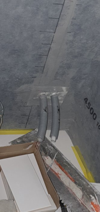 Två grå böjliga rör sticker ut från ett hål i en betongvägg, troligen del av VVS i badrum.