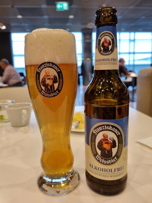 En skumtoppad weissbier i glas bredvid en flaska Franziskaner alkoholfri öl på bord.