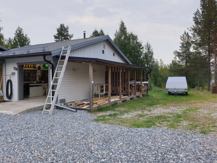 Garage med öppen port och arbetsbänkar, intilliggande vedförvaring och en släpvagn på grusplan.