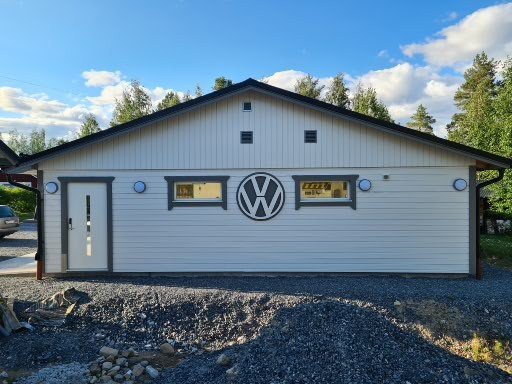 Vit garagebyggnad med 3 fönster, dörr och dekorativ VW-skylt ovanför garageporten, omgiven av grus.