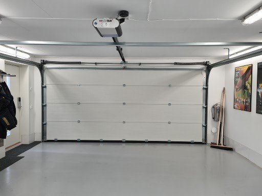 Interiör av ett nyligen färdigställt garage med slät, ljusgrå epoxymålat golv och vit rullport, LED-takbelysning och garageportöppnare.