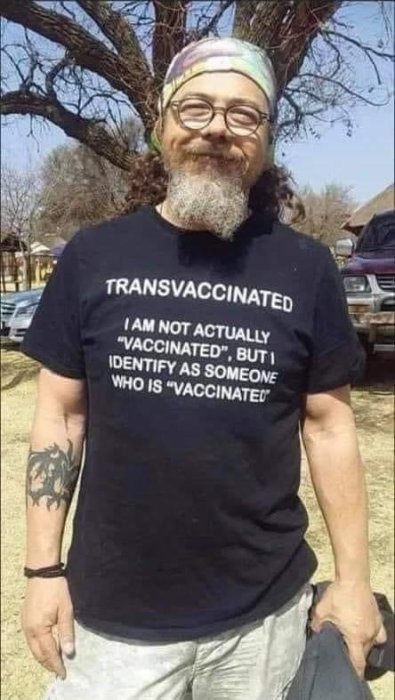 Person i svart T-shirt med texten "TRANSVACCINATED - ... IDENTIFY AS SOMEONE WHO IS 'VACCINATED'" och ett leende.