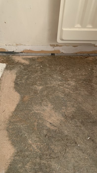 Dammtäckt golv vid vägghörna med misstänkt asbestludd, frågeställare osäker på materialet.