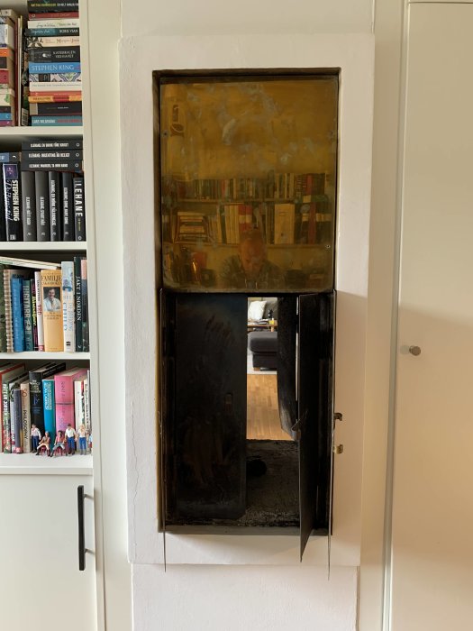 Inbyggd öppen spis med öppna svarta luckor i en vit murad vägg, speglar ett rum med bokhylla.