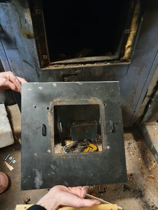 Metallplatta hålls upp framför öppningen av en panna där ved inkast luckan varit, med synliga verktyg och aska inuti.