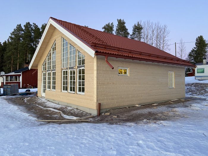 Nybyggd stomme till ett hus med taktegel och fönster på plats, omgivet av vintersnö och byggmaterial.