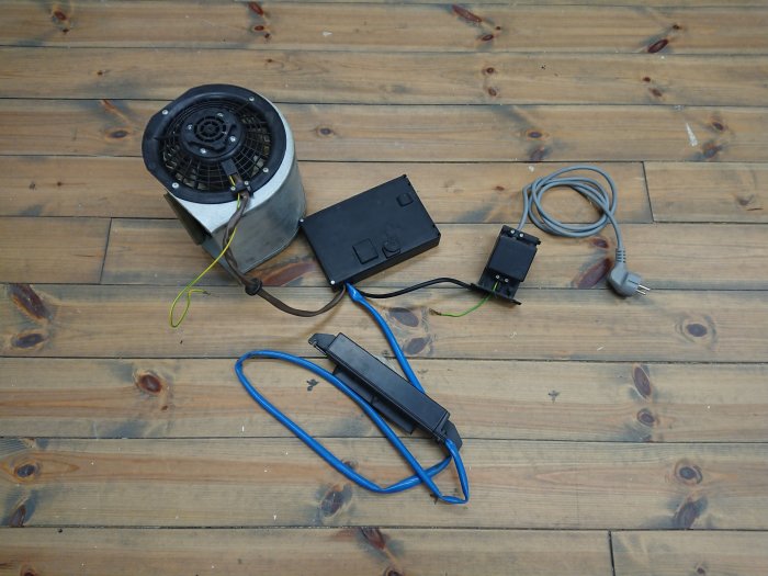 Komponenter till DIY luftrenare: fläktmotor, styrenhet med knappar, kablar och elkontakt på trägolv.