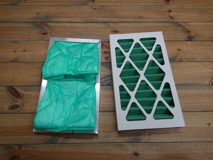 Gröna ersättningsfilter för luftrenare, oanvända och ligger på en trälaminatgolv, bredvid ett metallfiltergaller.