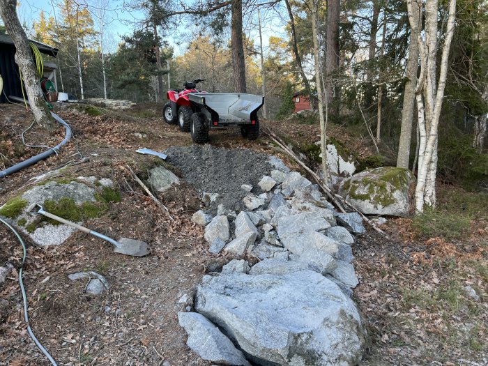 Nykonstruerad byggväg med spräckt sten på en skogstomt, med ATV och släpvagn i bakgrunden.