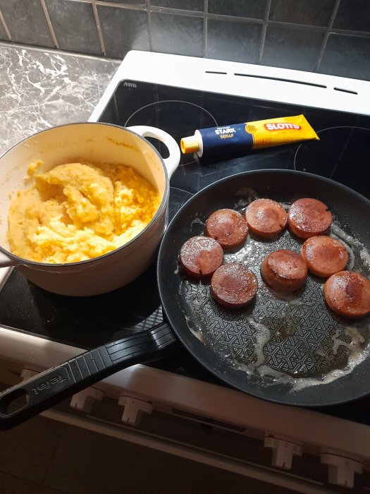 Stekt falukorv i panna och en kastrull med äggröra, tub med stark senap vid sidan på spishäll.